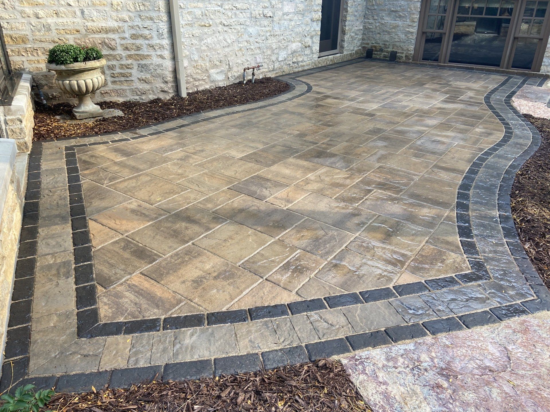 Patio with patterned pavers and dark border against stone wall, with mulch border and decorative pot.
