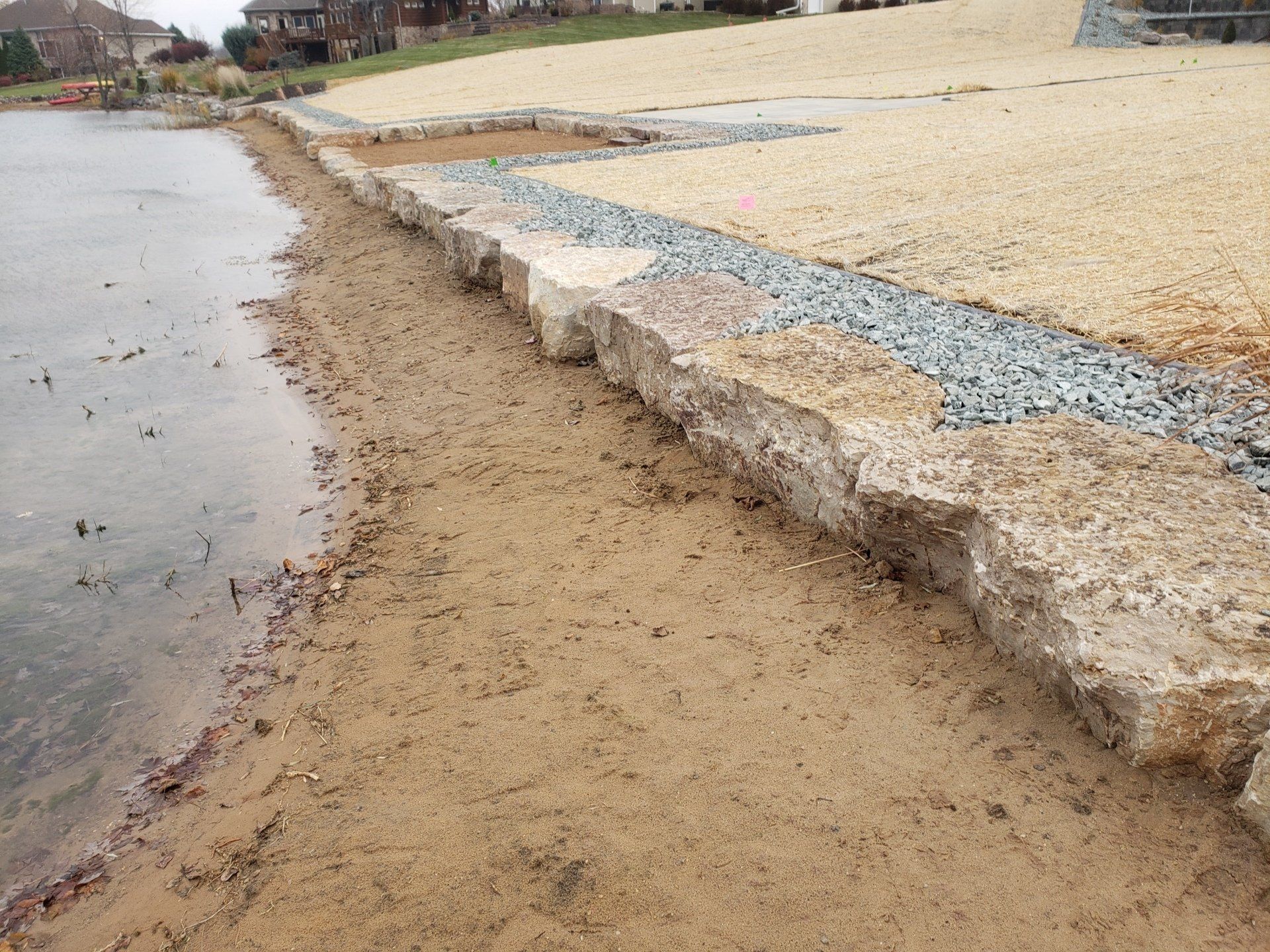 Stone retaining wall along a shoreline, with sand, gravel, and grass visible.