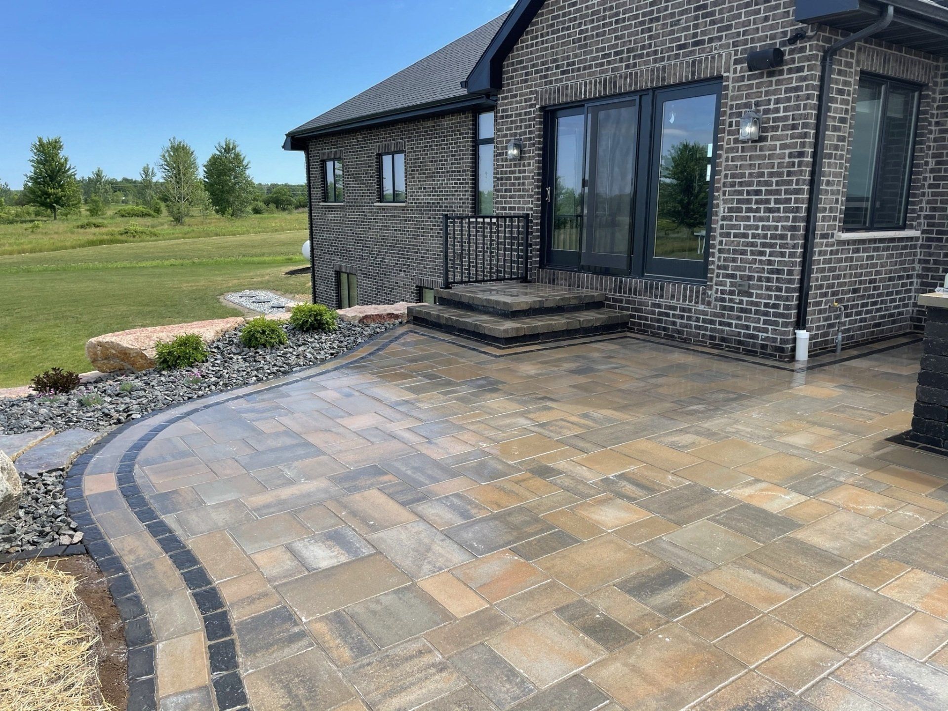 Brick house with a stone patio, steps, and black railing. Grass field in background.