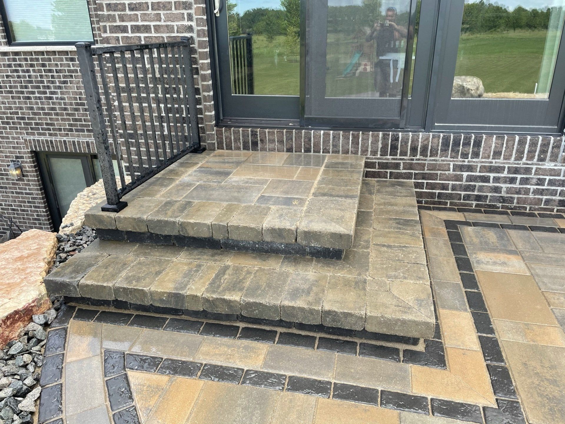 Brick steps leading to a glass door. A black metal railing is on the left. The ground is patterned with pavers.