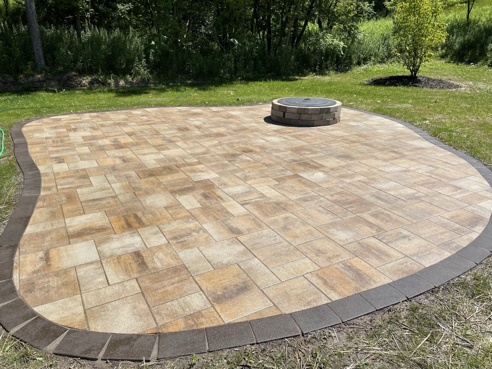 Stone paver patio with fire pit on a grassy lawn, bordered by darker pavers.