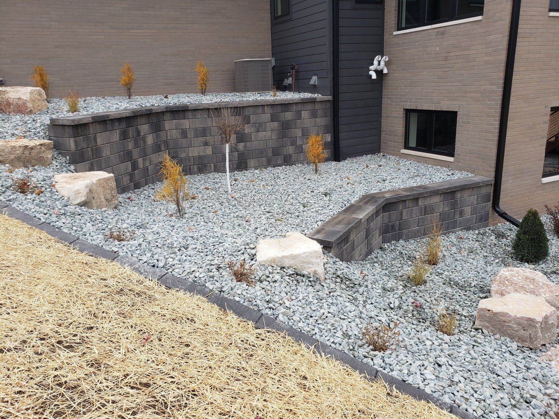 Landscaped yard with tiered retaining walls, rock beds, and small trees next to a building.
