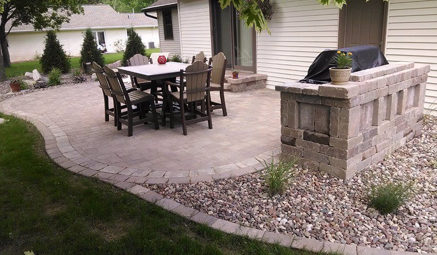 Patio with dining table, chairs, and built-in grill next to house; stone pavers and rock landscaping.