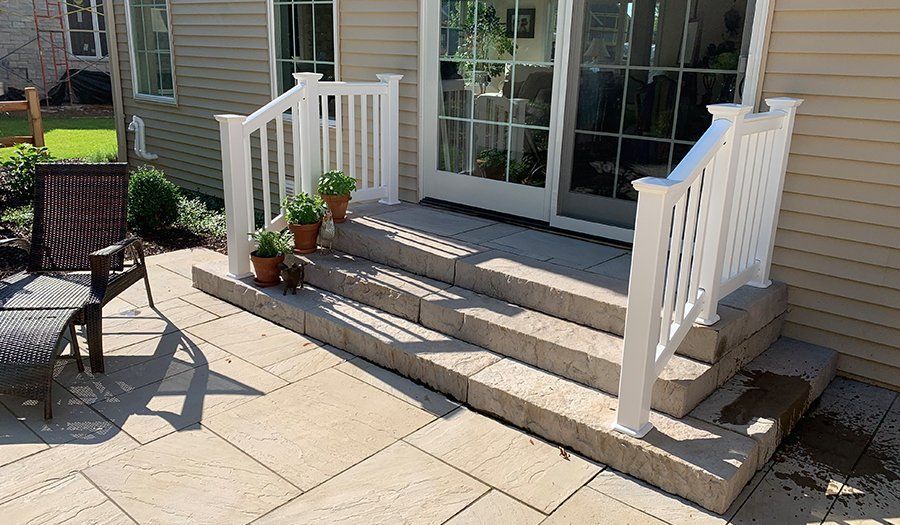 Patio steps leading to a sliding glass door. Beige pavers and white railing. Brown potted plants sit on top steps.