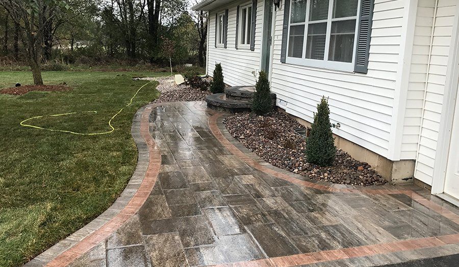 Pathway to a white house with wood-look pavers, red brick border, and small evergreens in a landscaped bed.