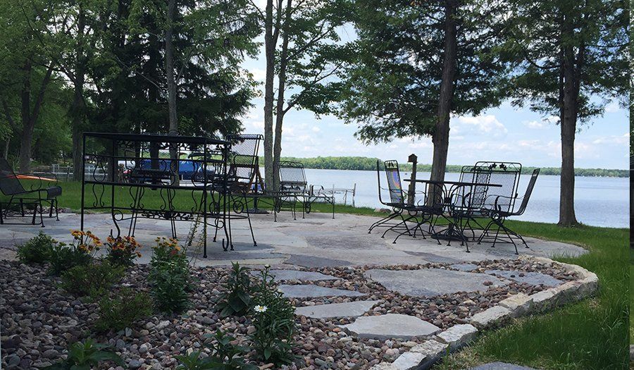 Patio overlooking lake with tables, chairs, and landscaping. Trees and water in the background.