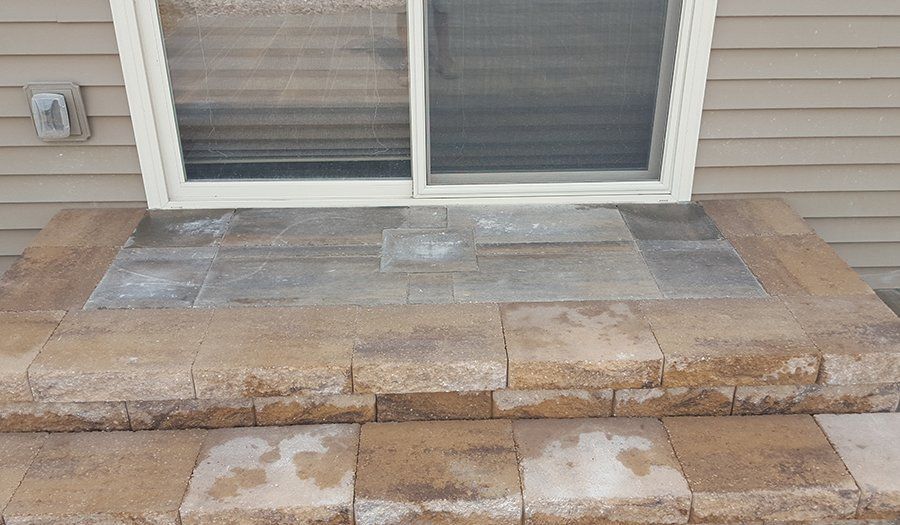 Two-step brick patio with a window above, against a tan house siding.