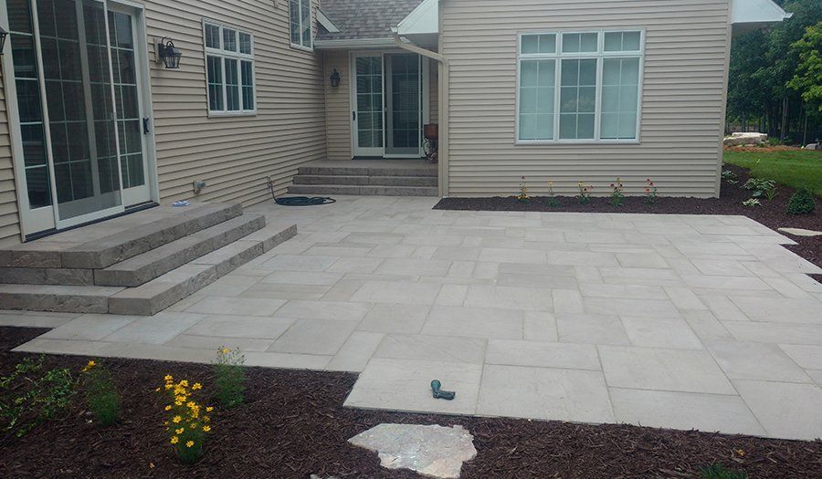 Backyard patio with light gray stone tiles, steps, and landscaping beds with dark mulch and plants.