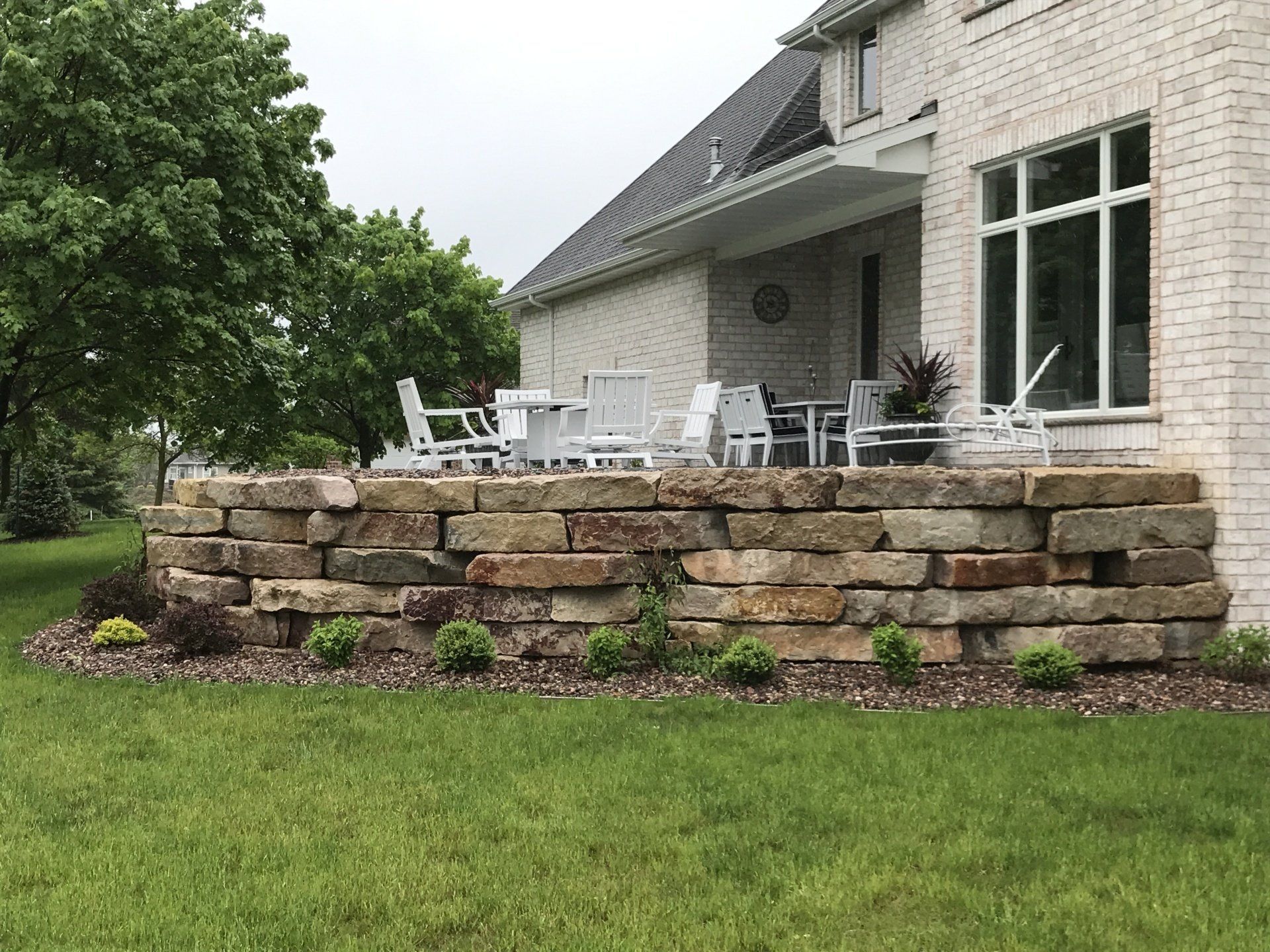 Stone retaining wall supports patio with white furniture, beside brick house. Green lawn.