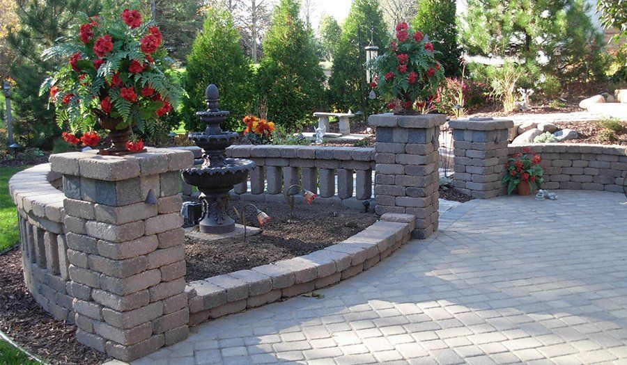 Stone patio with fountain, flower planters, and brick walls in a sunny garden.