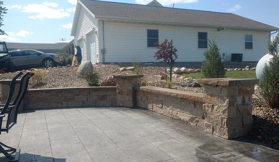 Stone patio with a low retaining wall, plants, and a white house with a garage in the background.