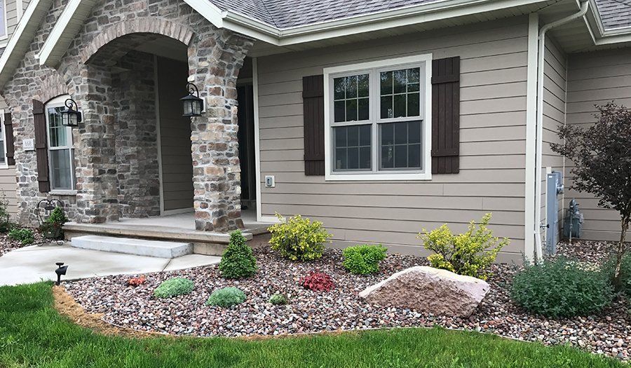 Tan house with stone archway, brown shutters, and landscaped front yard with rocks and plants.
