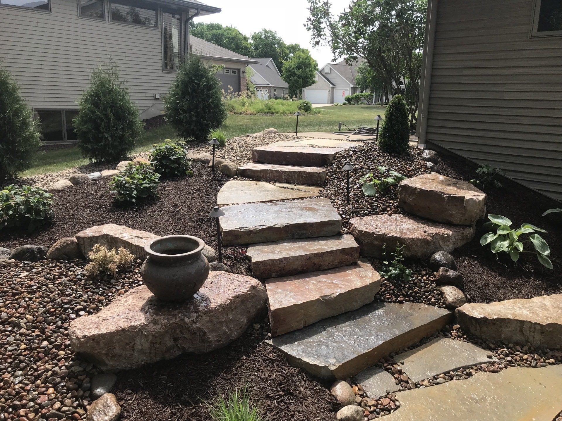 Stone steps lead through a landscaped yard, with boulders, mulch, and plants surrounding.