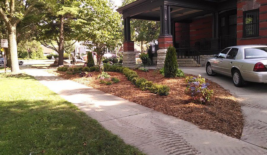 A landscaped front yard with a driveway, sidewalk, and a parked car next to a brick house with a porch.