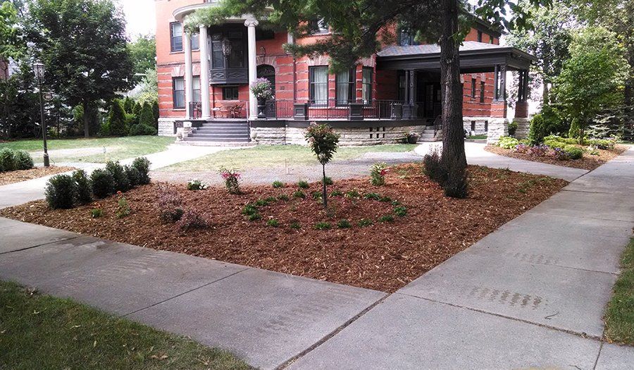 Brick house with a landscaped yard and trees, bordered by a sidewalk.