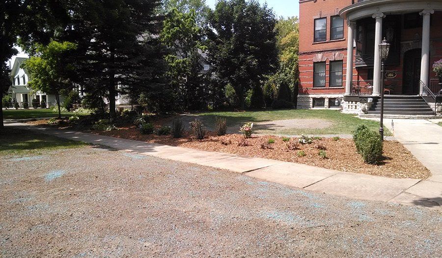 Gravel drive curves past a mulched garden bed and sidewalk toward a large brick building with columns.