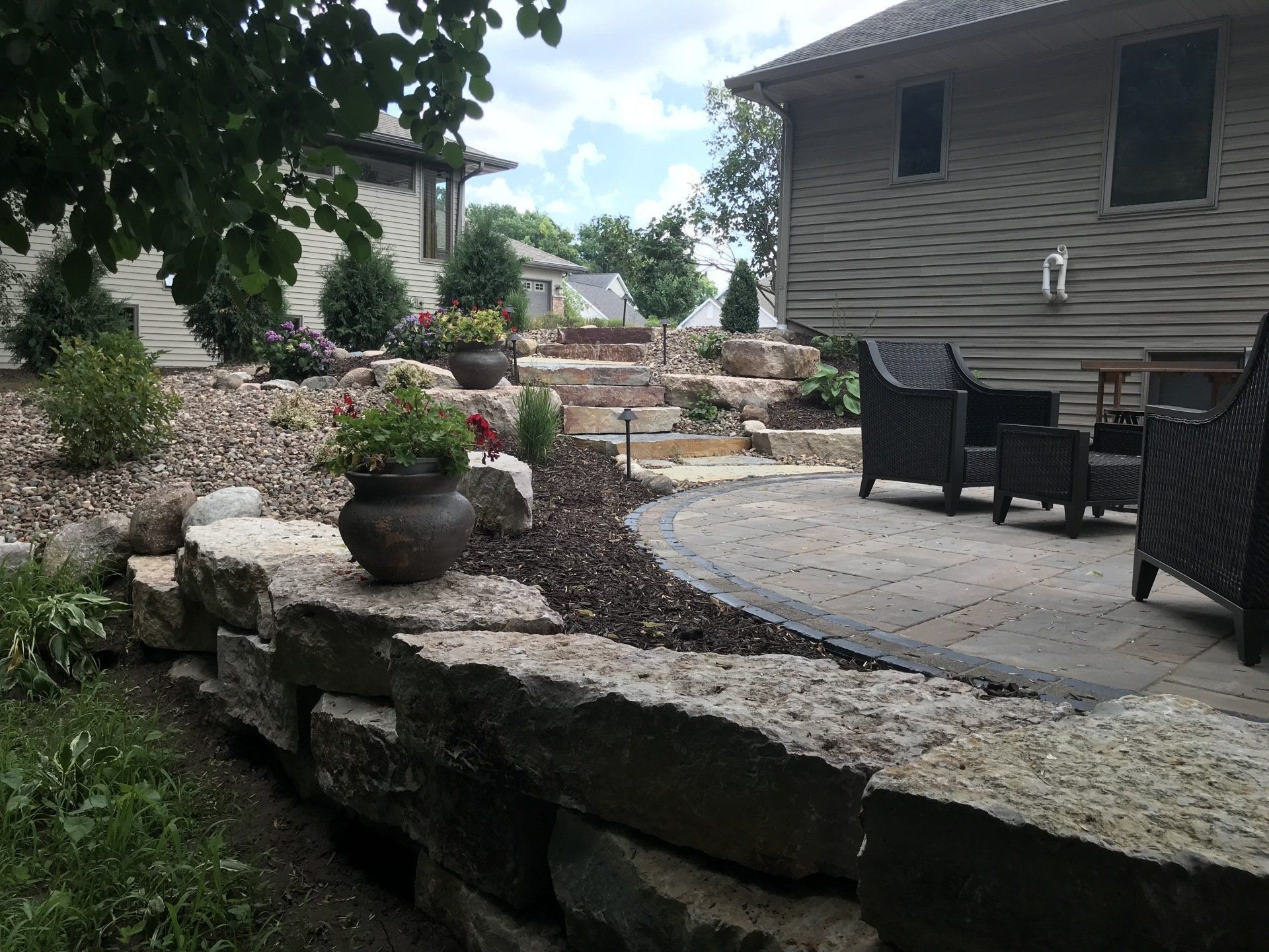 Stone retaining wall and steps lead to a patio with wicker furniture. Landscaped backyard with flowers and greenery.