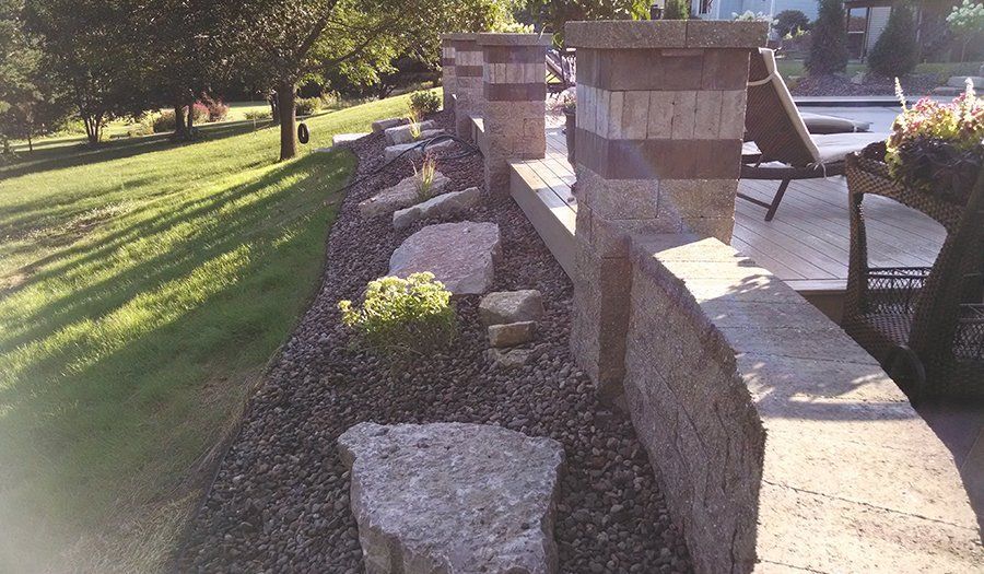 Retaining wall with stone pillars and landscaping along a patio. Green lawn and trees in background.
