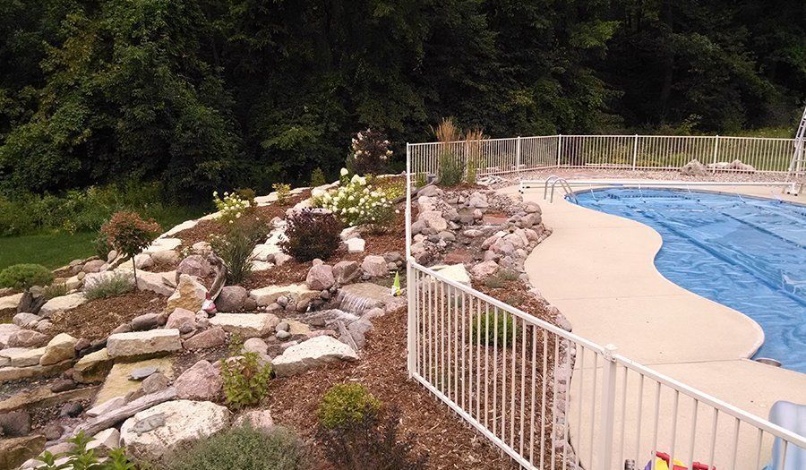 Pool area with white fence, landscaping with rocks and plants, trees in the background.