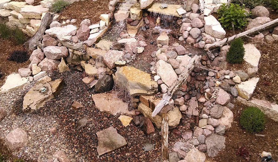 Rock garden with waterfall feature, brown soil, gray and tan rocks, small plants.