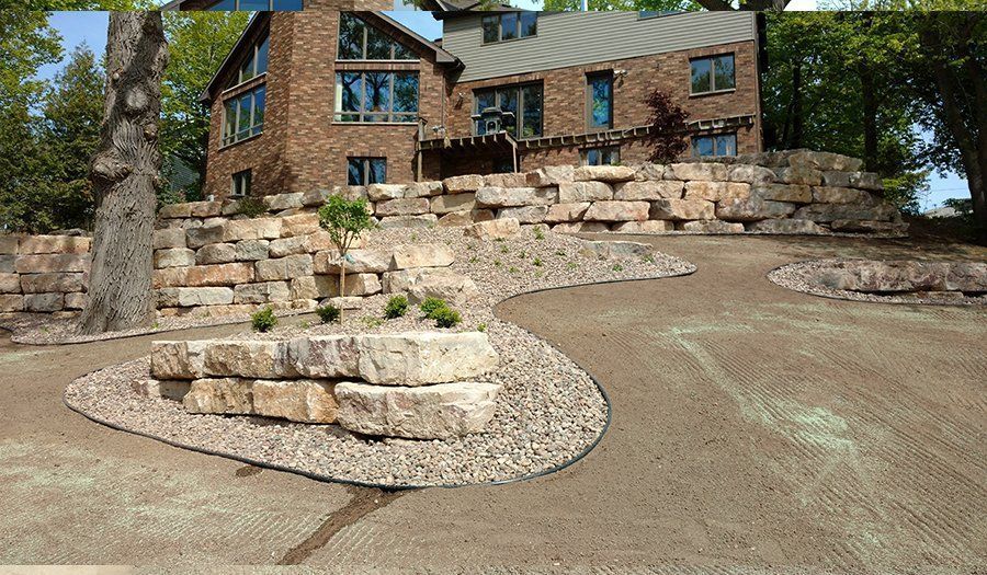 Stone retaining walls and gravel path leading to a two-story brick house with large windows, landscaping.