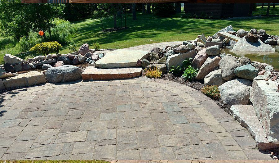 Stone patio with steps leading to a pond surrounded by large rocks and greenery.
