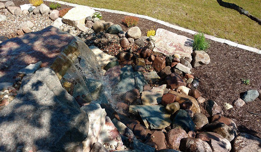 Water flows down rocks into a small stream, part of a landscaped garden with grass and mulch.