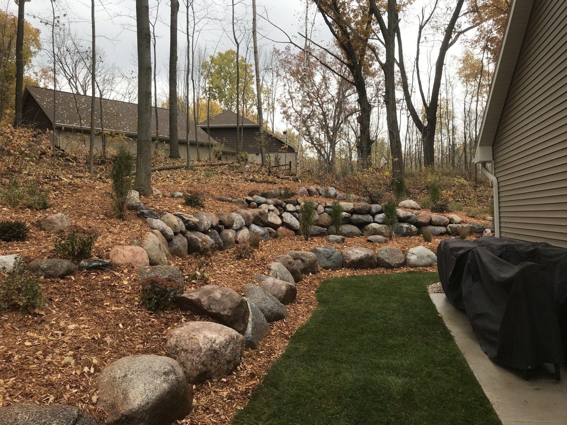 Backyard with stone retaining walls, grassy area, and trees; house in background.