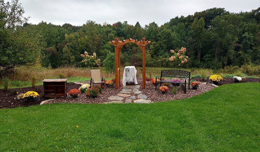 Arched wooden arbor with seating area, flowers, and bench in front of a wooded backdrop on a grassy lawn.