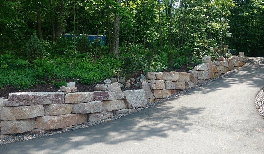 Stone retaining wall along a paved driveway, with trees and greenery in the background.
