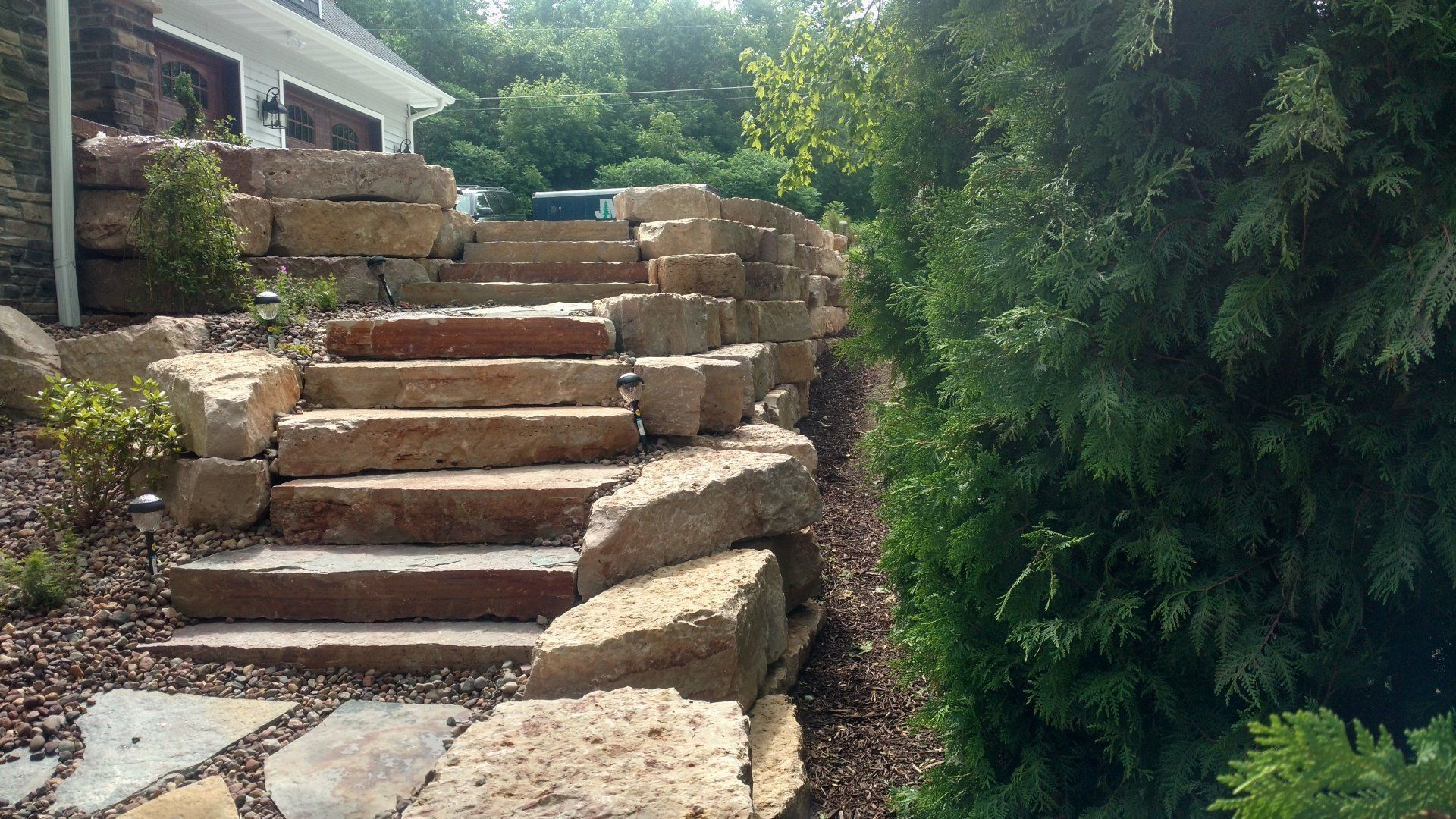 Stone steps leading uphill, alongside a house and greenery, with a bright sky.