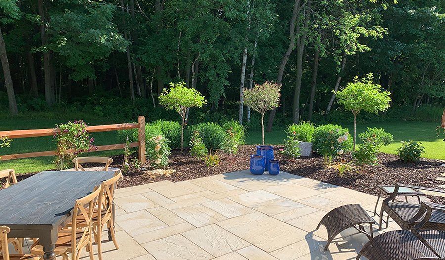 Patio with stone pavers, wooden furniture, and garden with trimmed trees and shrubs against a forested backdrop.