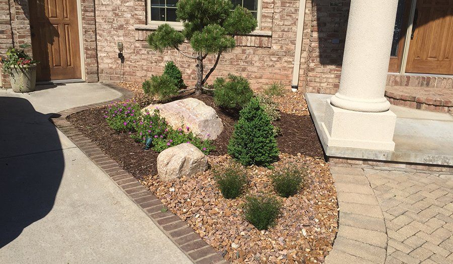 Landscaped flower bed with rocks, mulch, and various green plants in front of a brick house.