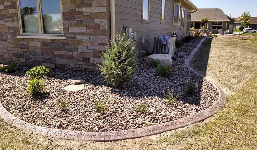 Landscaped flower bed with stone edging, brown gravel, and plants next to a house.