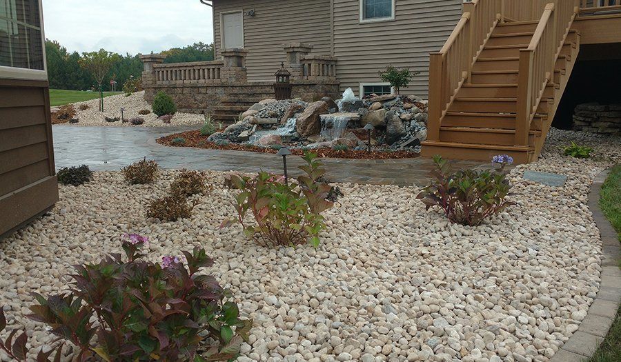 Landscaped yard with white rocks, plants, and a waterfall feature near a deck and house.