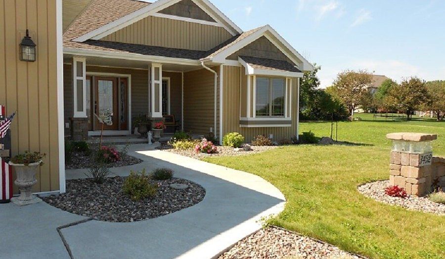 Tan house with concrete walkway, green lawn, and stone landscaping. Bright sunny day.