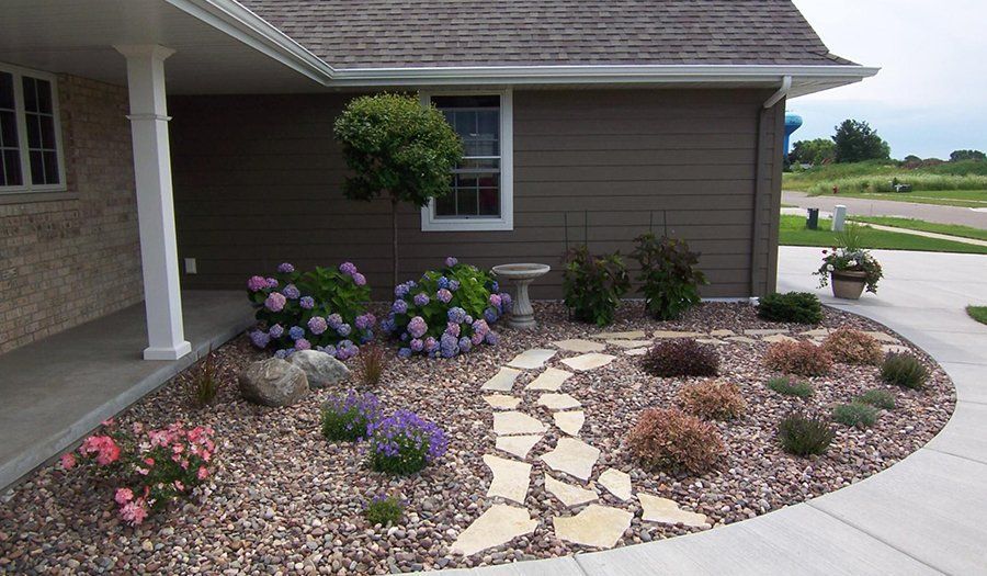 Landscaped garden bed with stone path, flowers, and shrubs in front of a house.