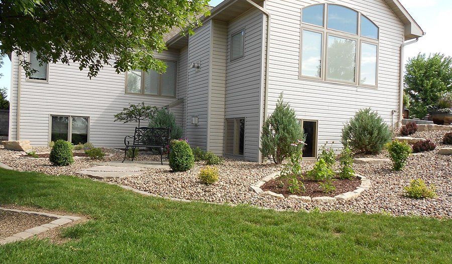 Two-story house with light siding and large arched window. Landscaped yard with gravel, shrubs, and green lawn.