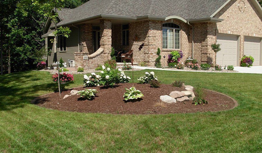 Brick house with circular flower bed of white and green plants surrounded by brown mulch, neat green lawn.