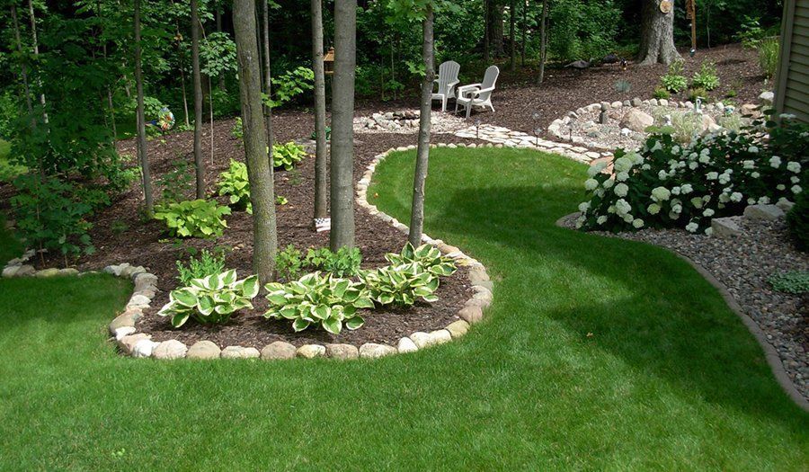 Landscaped yard with green grass, trees, and flower beds edged with stones.
