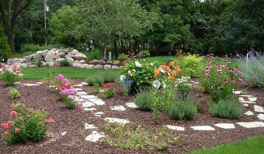 Flower garden with stone path, various colorful flowers, green bushes, and trees in the background.