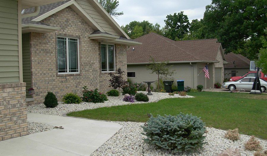 House with beige brick facade, landscaped yard with gravel, grass, and shrubs. A sidewalk leads to the entrance.