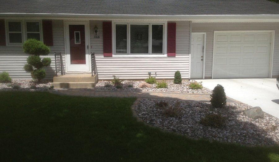 A gray house with maroon shutters and a red door, featuring a landscaped front yard with shrubs and a concrete driveway.