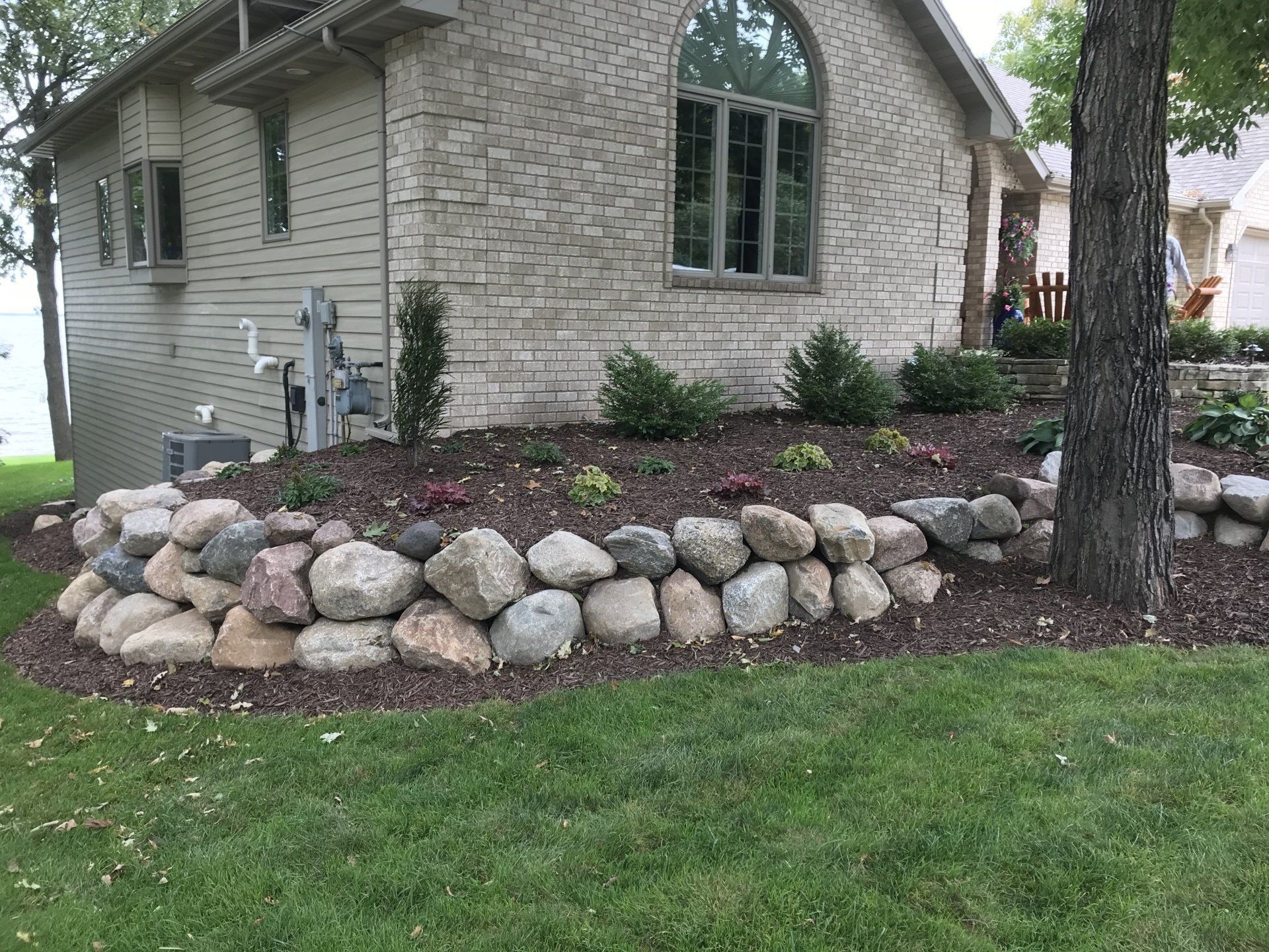 Rock retaining wall in front of a light brick house with a green lawn.