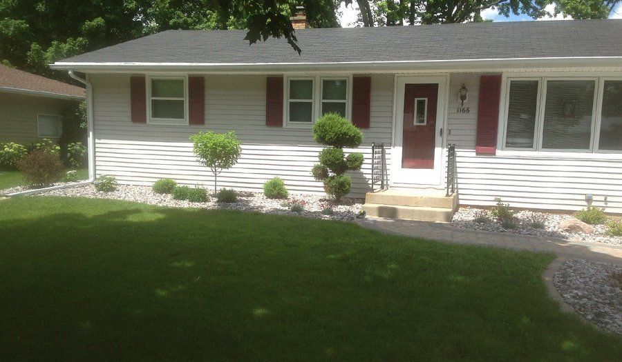 Tan house with red shutters and door, landscaping with grass and stone.