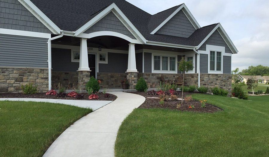 Gray house with stone and shingle siding, white columns, and a concrete path. Lush green lawn and garden.
