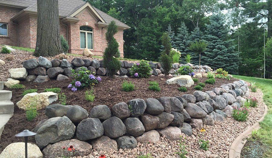 Stone retaining walls frame a landscaped yard in front of a brick house, featuring trees, flowers, and a grassy area.