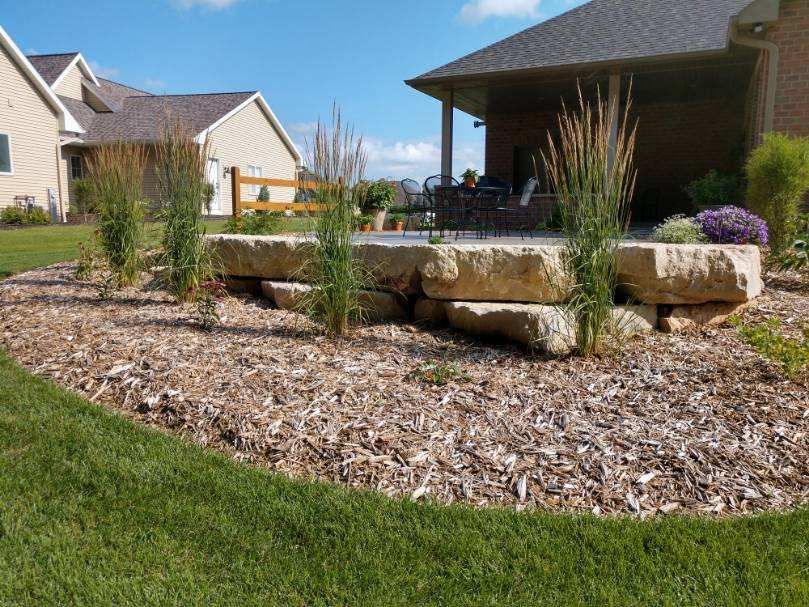 Stone patio with tall grasses, surrounded by mulch and green lawn, next to a house.