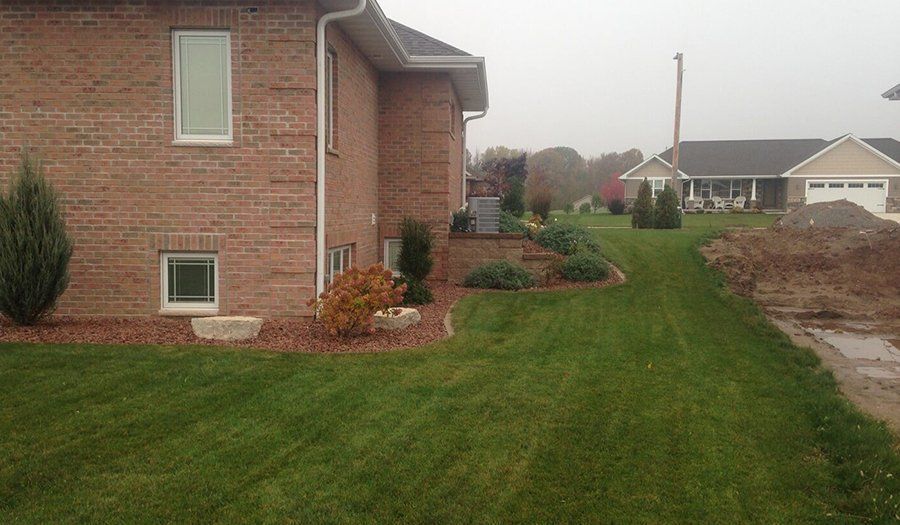 Brick house with a green lawn, garden bed, and a neighbor's house in the background on a cloudy day.
