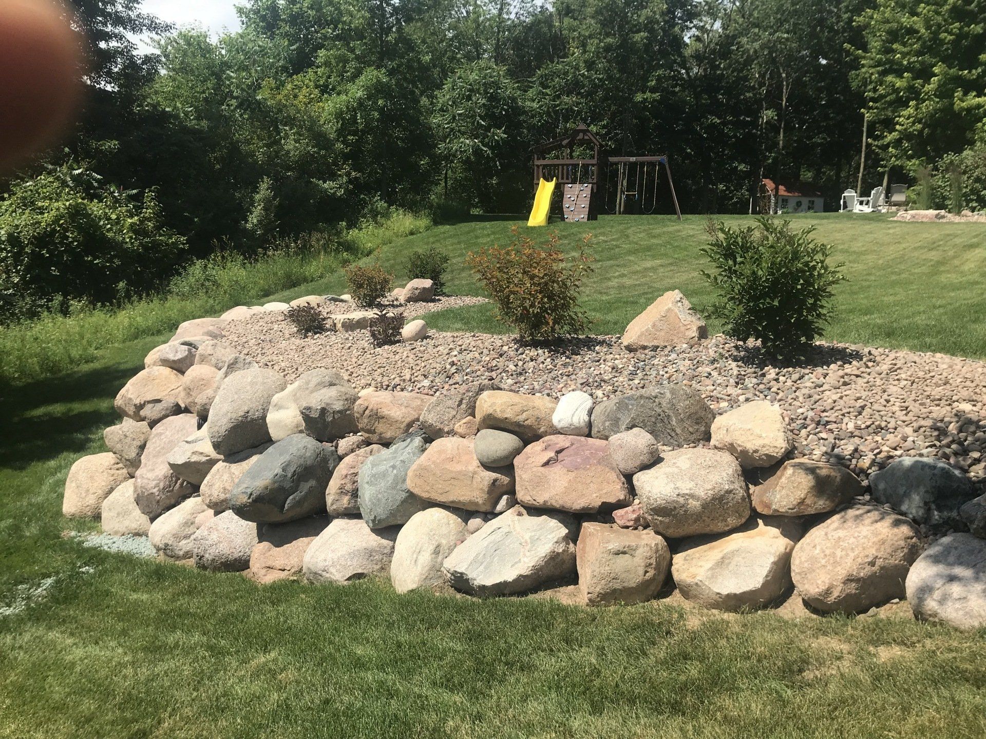 Rock retaining wall with bushes and gravel in a grassy yard, with a playset in the background.